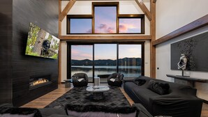 A modern living room with a fireplace, large windows, and a view of a lake. - LAKEFRONT - 5 STAR 4,500 sqft! Hot Tub, Fire Pit, Dock, Fireworks & Fireworks (Big Bear Lake)