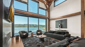 A spacious living area with a large window overlooking a body of water, a fireplace, and modern furniture. - LAKEFRONT - 5 STAR 4,500 sqft! Hot Tub, Fire Pit, Dock, Fireworks & Fireworks (Big Bear Lake)