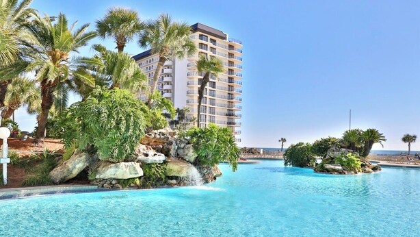 A resort pool with clear blue water, surrounded by palm trees and a large building in the background. - Sweeping Gulf Views! 1/2 sleeps 4! (Panama City Beach)