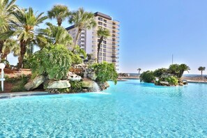 A resort pool with clear blue water, surrounded by palm trees and a large building in the background. - Sweeping Gulf Views! 1/2 sleeps 4! (Panama City Beach)