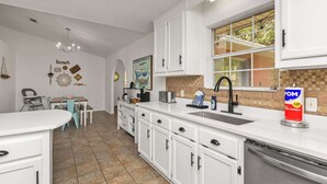 Una cocina con armarios blancos, un lavamanos y una ventana. Hay un cuadro en la pared y una silla en el comedor.