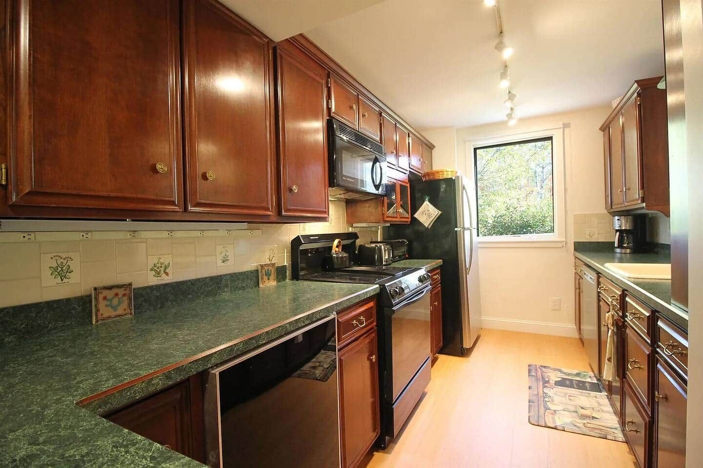 A kitchen with wooden cabinets, a stove, a refrigerator, and a window with a view of greenery.