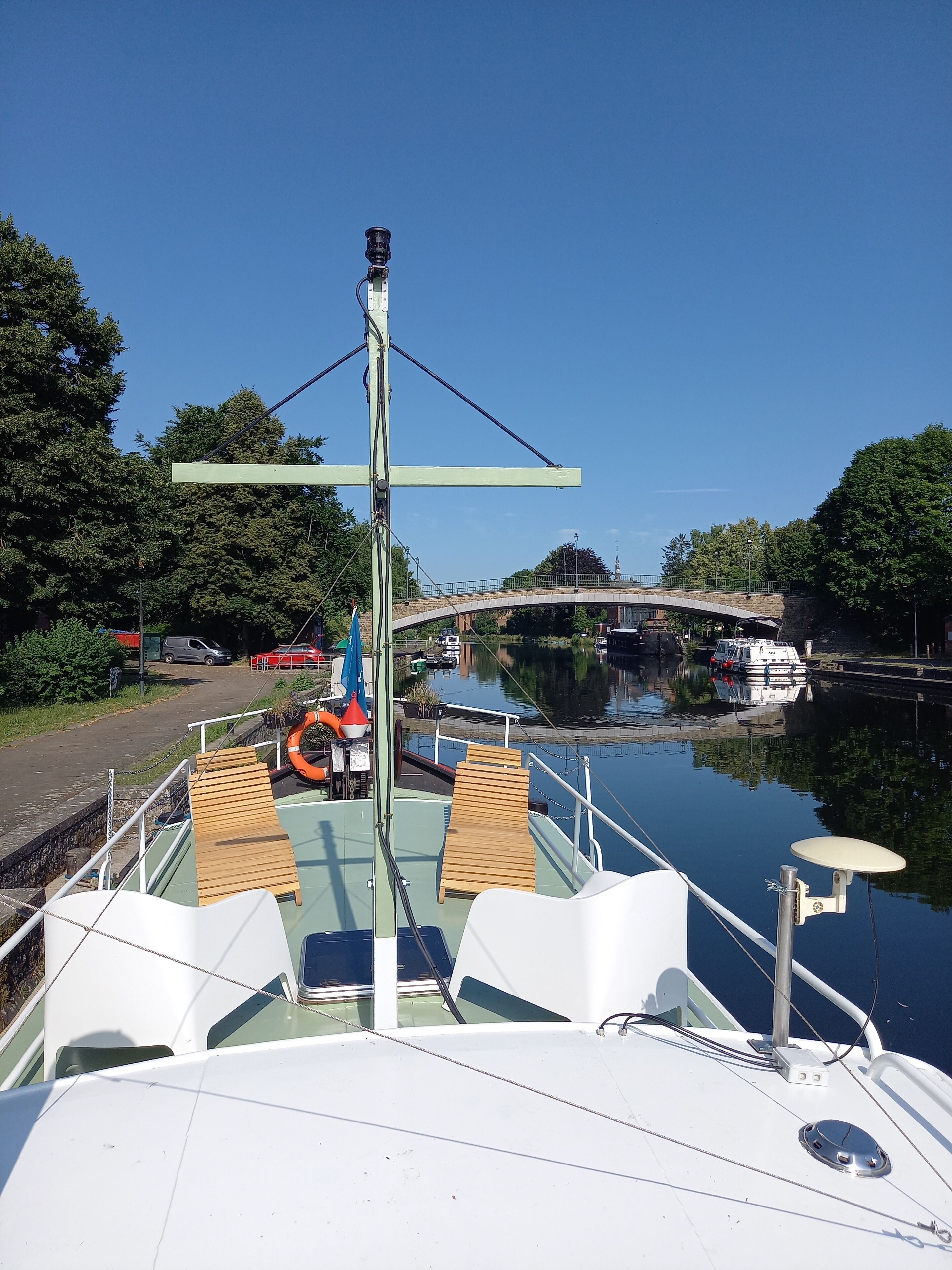 Un bote con cubierta de madera, barandales de metal y un flotador salvavidas. Se observa un puente al fondo.