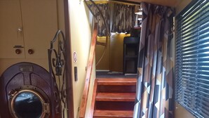 Eine Holztreppe führt zu einer Zwischenebene mit einem gemusterten Vorhang und einer Tür mit rundem Fenster.