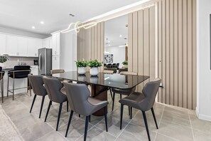 A modern kitchen with white cabinets, a stainless steel refrigerator, and a dining area with a glass table and grey chairs. - 5BR house with game room near Disney area (Kissimmee)