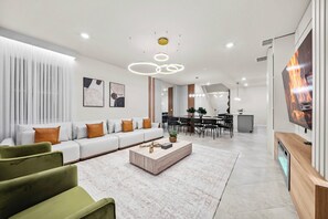 A modern living room with a sofa, coffee table, and green armchairs. There is a dining area in the background with a TV mounted on the wall. - 7BR house sleeps 20 with game room (Clermont)