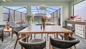 A dining area with a wooden table and chairs, a hanging light fixture, and a view of a swimming pool through a glass enclosure. - 7BR house sleeps 20 with pool table (Clermont)