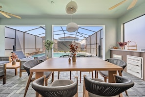 A dining area with a wooden table and chairs, a hanging light fixture, and a view of a swimming pool through a glass enclosure. - 7BR house sleeps 20 with pool table (Clermont)