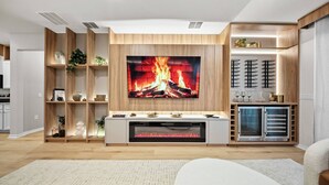 A modern living room with a large flat-screen TV mounted on a wooden wall, a built-in fireplace below it, and a white sofa in the foreground. - 7BR house sleeps 20 with pool table (Clermont)