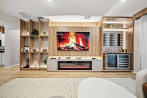 A modern living room with a large flat-screen TV mounted on a wooden wall, a built-in fireplace below it, and a white sofa in the foreground. - 7BR house sleeps 20 with pool table (Clermont)