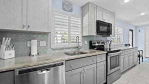 A modern kitchen with stainless steel appliances, white cabinetry, and a large window with blinds. - Longboat Key - Choose Joy - Relax on the Beach (Longboat Key)