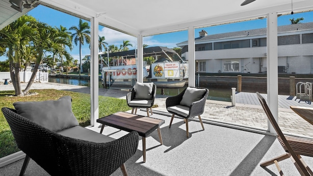 A covered outdoor seating area with wicker chairs and a table, overlooking a canal with a boat and buildings in the background. - Longboat Key - Choose Joy - Relax on the Beach (Longboat Key)