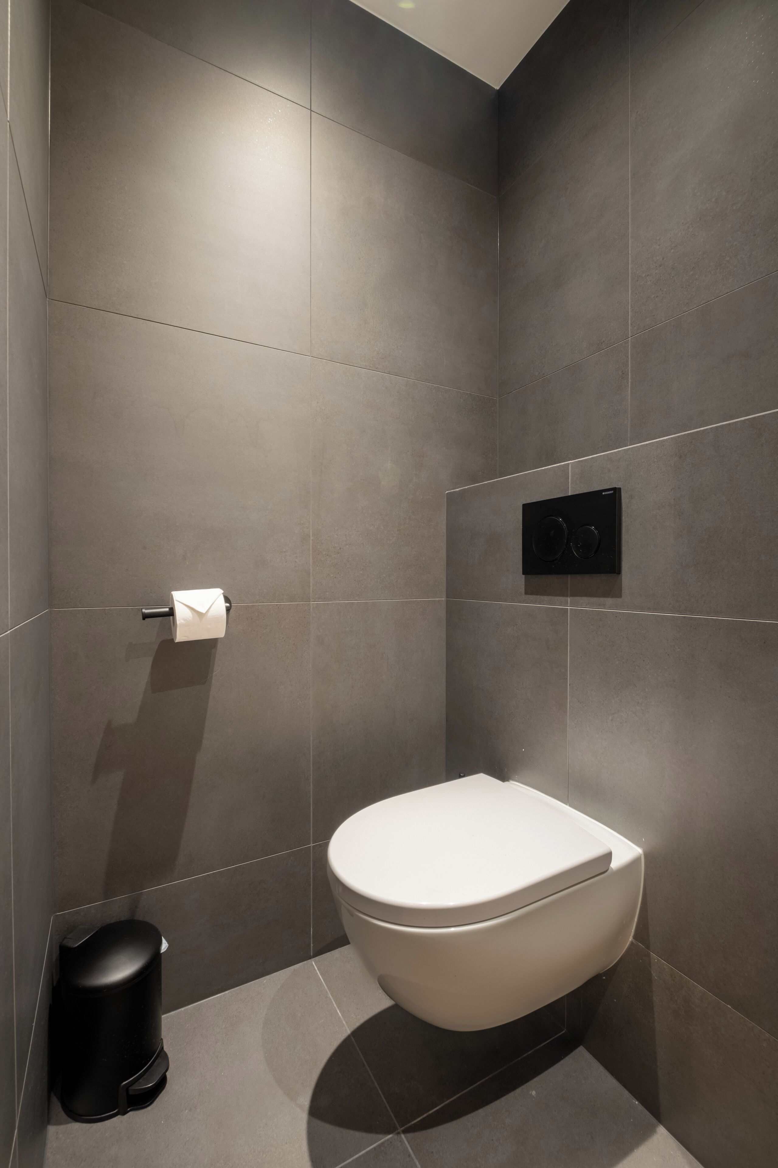 Een moderne badkamer met een wit toilet, een zwarte prullenbak en een papieren handdoekdispenser aan de muur.