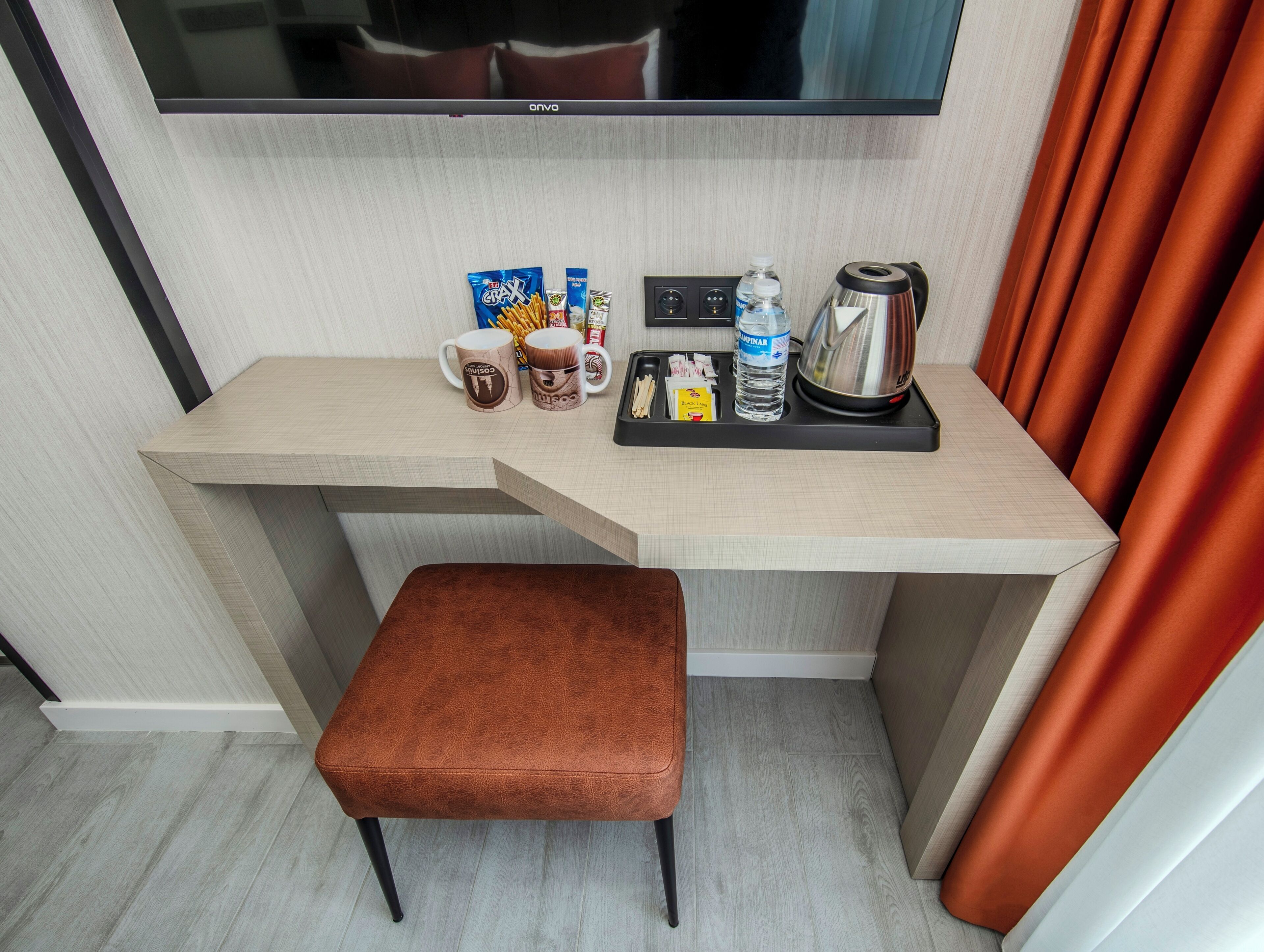Une chambre d’hôtel dotée d’un bureau en bois, d’un banc rembourré de couleur marron, d’un téléviseur à écran plat et d’un mini-bar contenant des en-cas et une bouilloire.
