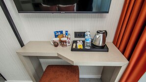 A hotel room with a wooden desk, a brown cushioned bench, a flat-screen TV, and a minibar with snacks and a kettle. - Cosinüs Airport Hotel (Arnavutköy)