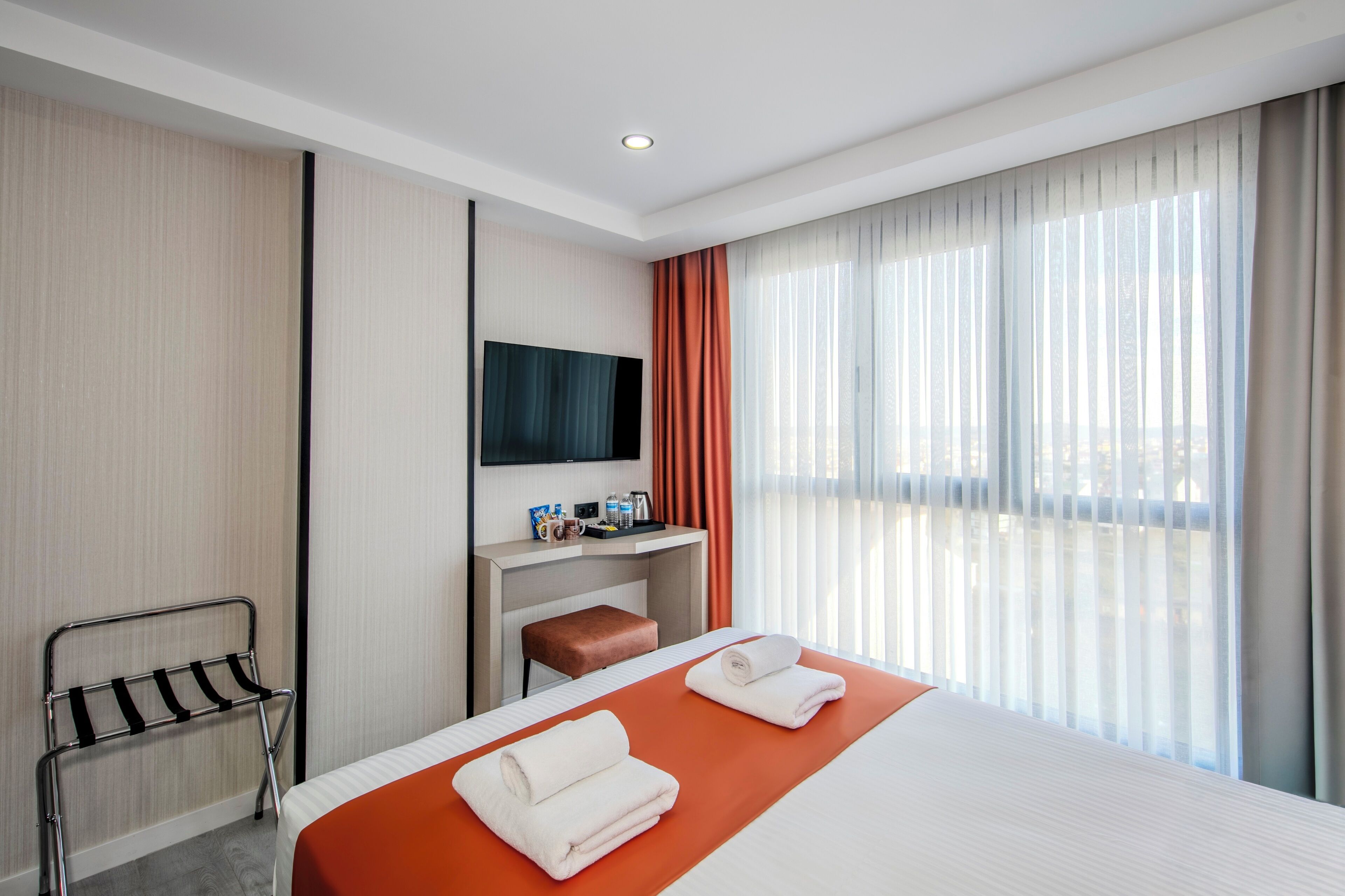 Une chambre d’hôtel avec un lit, une télévision, une petite table avec des articles de toilette, un banc et une fenêtre avec des rideaux.