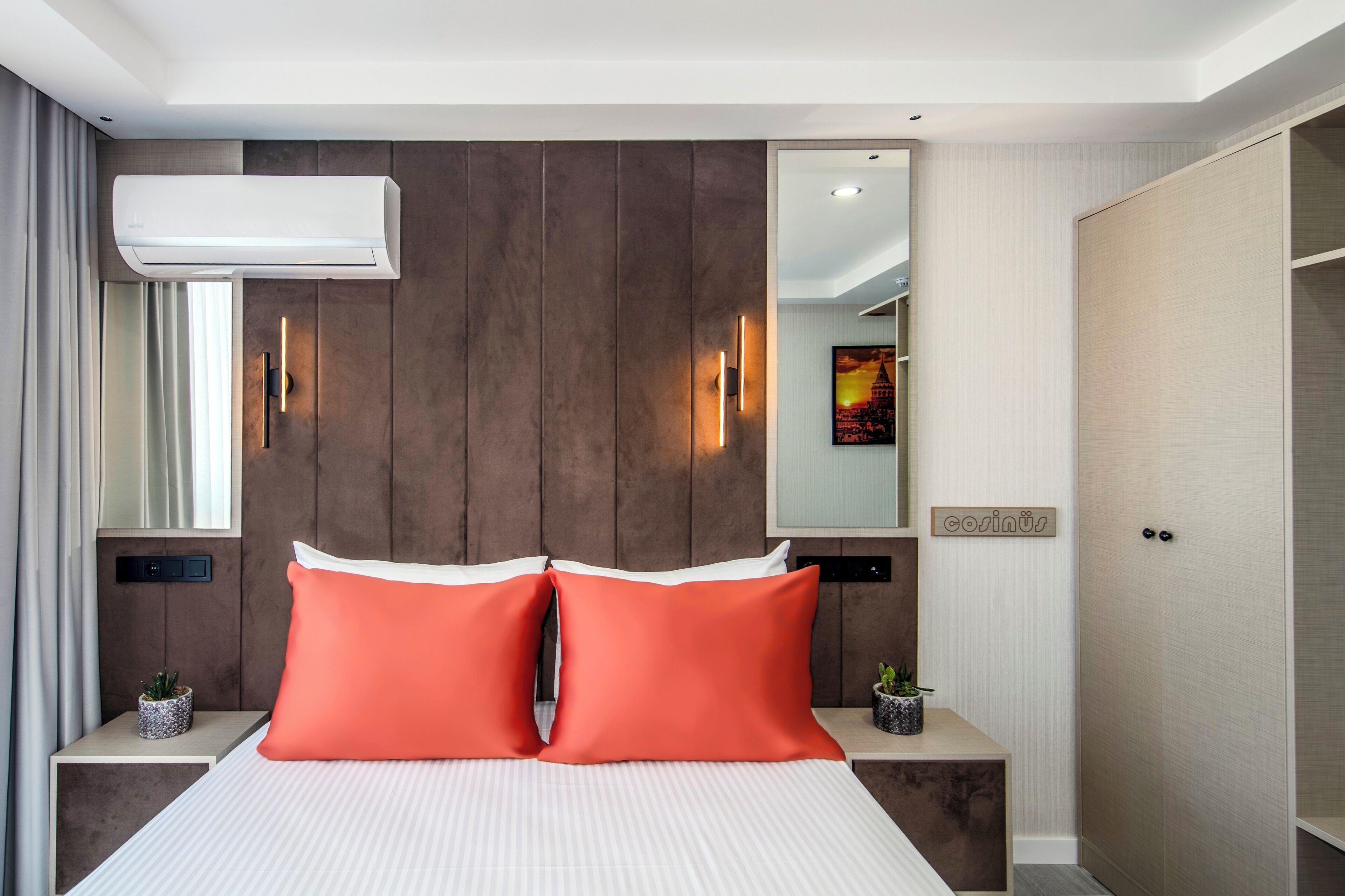 Una habitación de hotel con cama, dos mesitas de noche, aire acondicionado, espejo, luz en la pared y una puerta con manija.