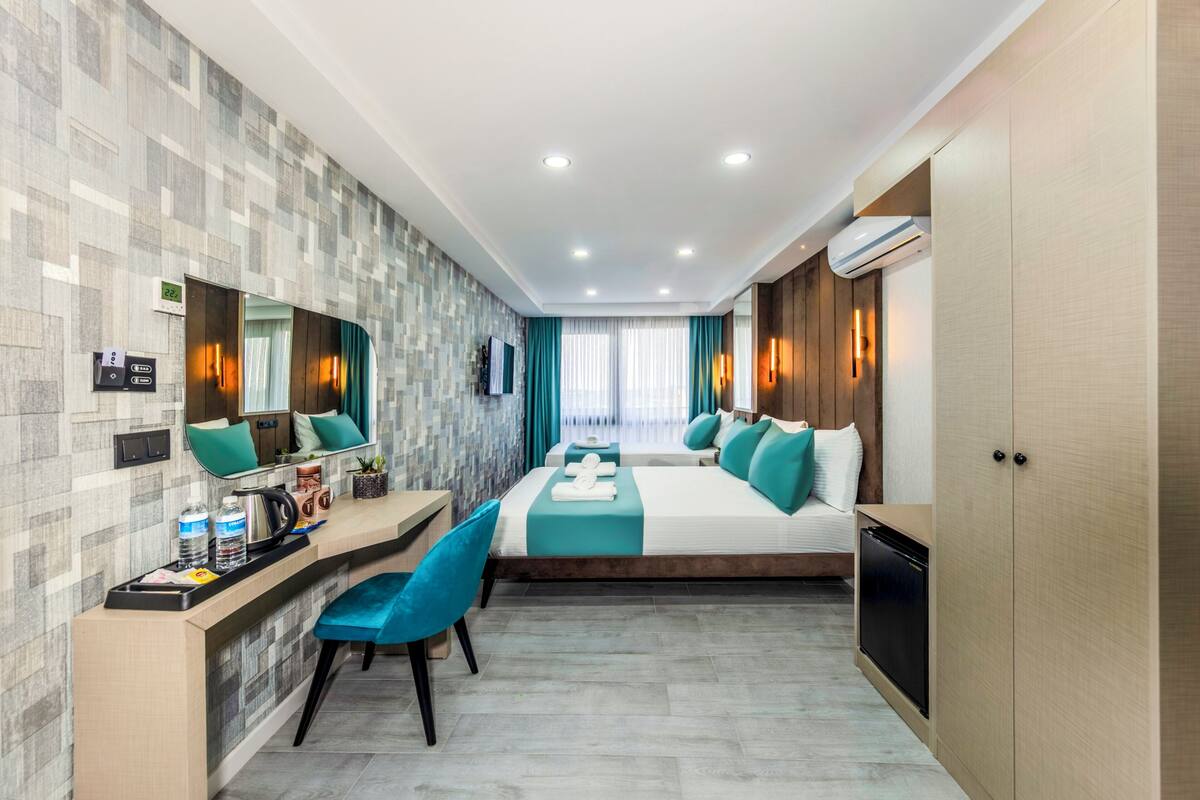 Quarto de hotel moderno com uma cama grande, uma mesa de trabalho com cadeira, uma pequena cozinha e um banheiro com chuveiro e vaso sanitário.