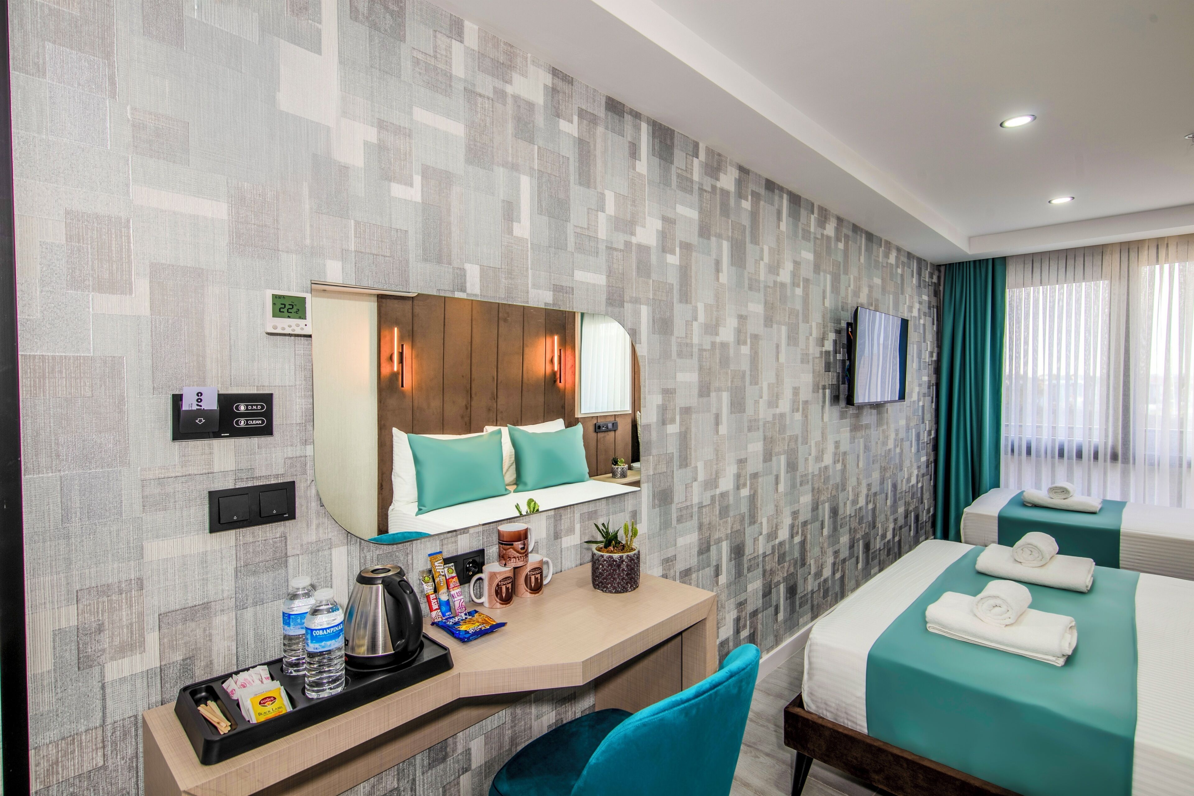 A hotel room with a modern design, featuring a large bed with teal bedding, a desk with a coffee maker, and a small plant.