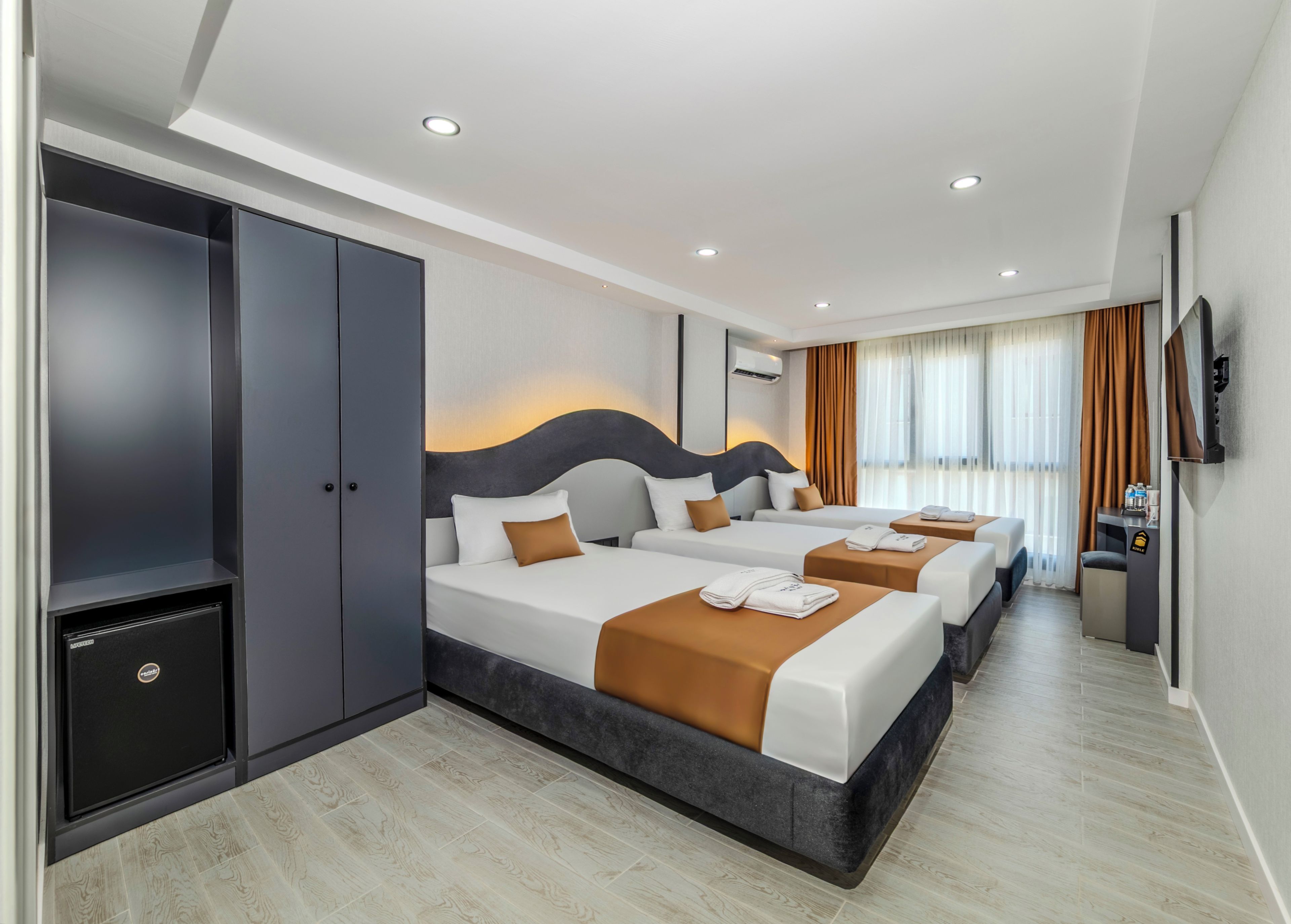 Habitación de hotel con un armario grande, dos camas con ropa de cama marrón y blanca, un televisor de pantalla plana y un minibar.