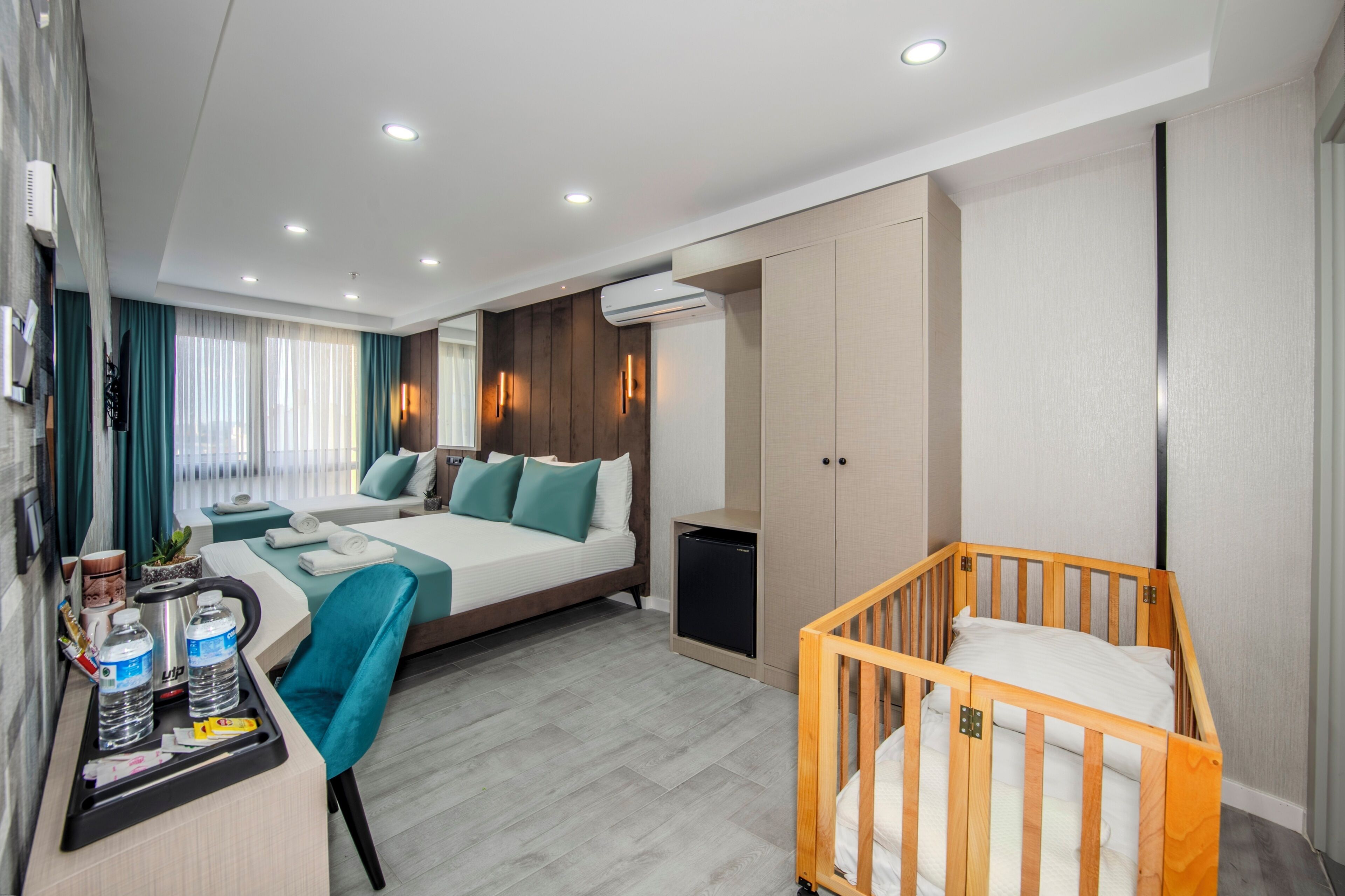 A modern hotel room with a large bed, a desk with amenities, a crib, and an air conditioning unit.