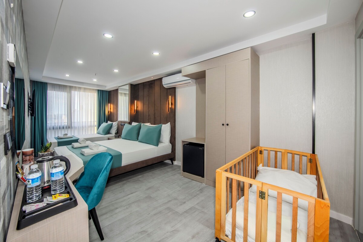 A modern hotel room with a large bed, a desk with amenities, a crib, and an air conditioning unit.