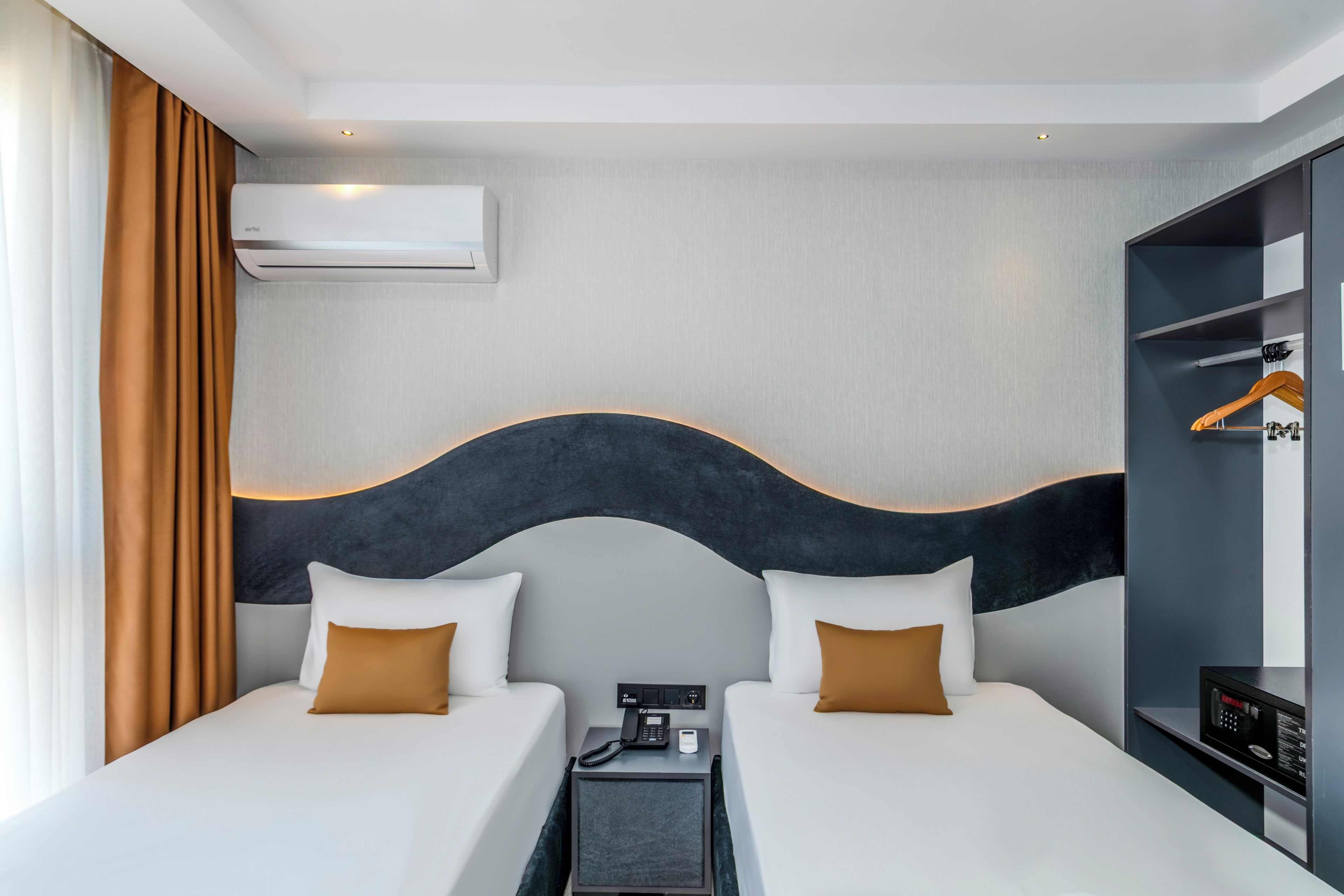 A hotel room with two beds, a wardrobe, a bedside table with a phone, and an air conditioning unit.