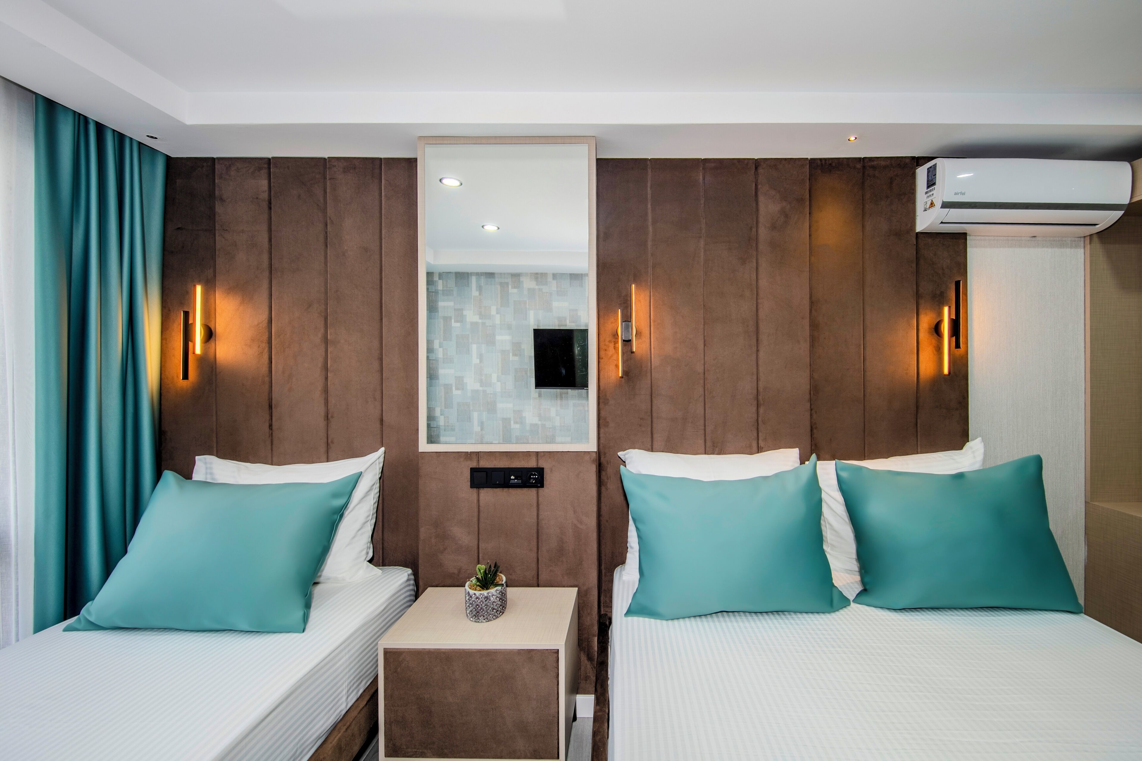 A hotel room with two beds, teal pillows, a small bedside table with a plant, and an air conditioning unit.