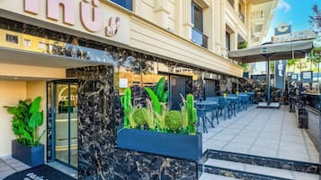 Entrada de hotel con diseño moderno, que incluye un mostrador de recepción, una zona de estar con mesas y sillas, y una gran maceta con plantas.