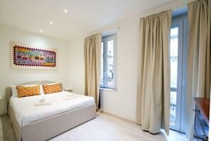 Un dormitorio con cama, cortinas color crema, una ventana y una obra de arte colorida en la pared.