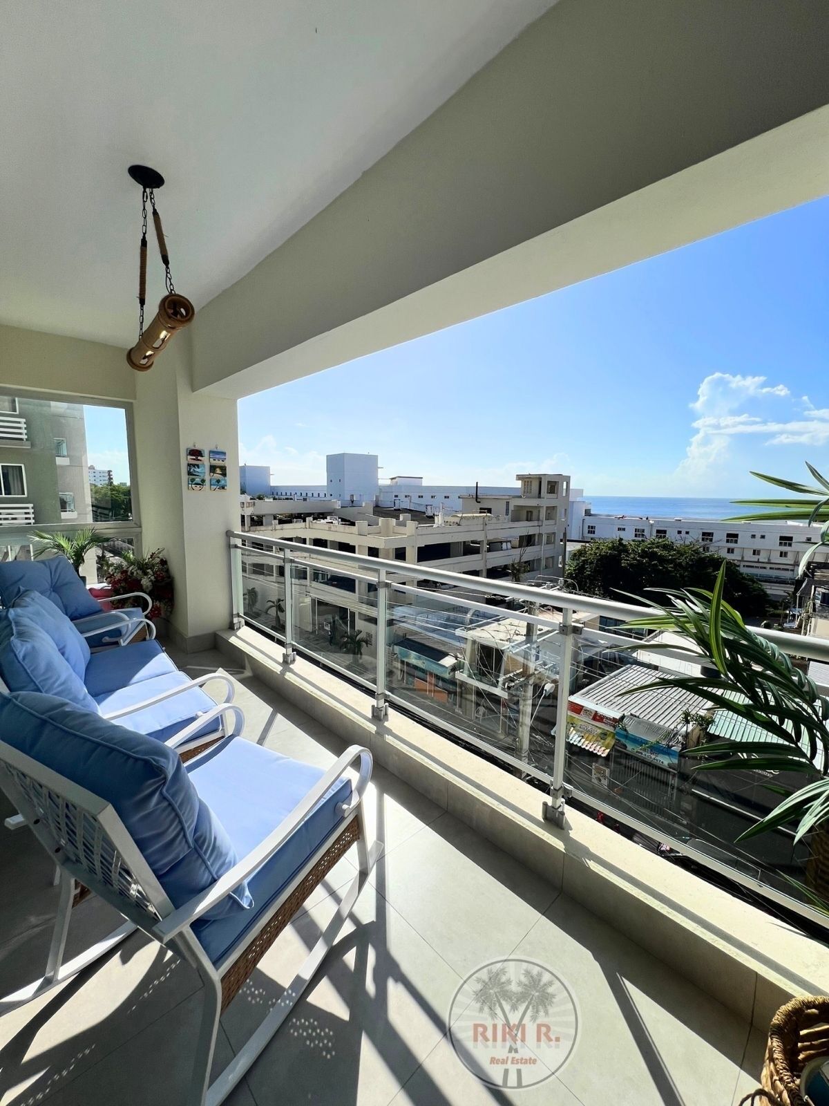 A balcony with blue lounge chairs, a hanging light fixture, and a view of buildings and the sea.
