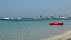 En strand med klart vand, en rød båd og industrielle bygninger i baggrunden.