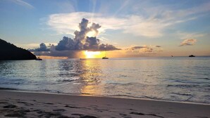 Una playa con un atardecer, mar tranquilo y barcos.
