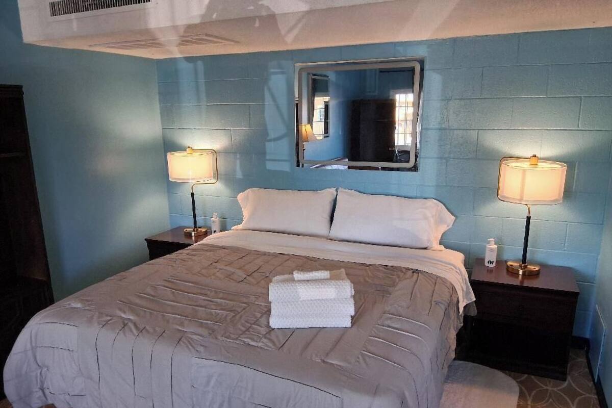 Une chambre d’hôtel avec un grand lit, deux tables de chevet avec des lampes, un miroir au-dessus du lit et un mur bleu.