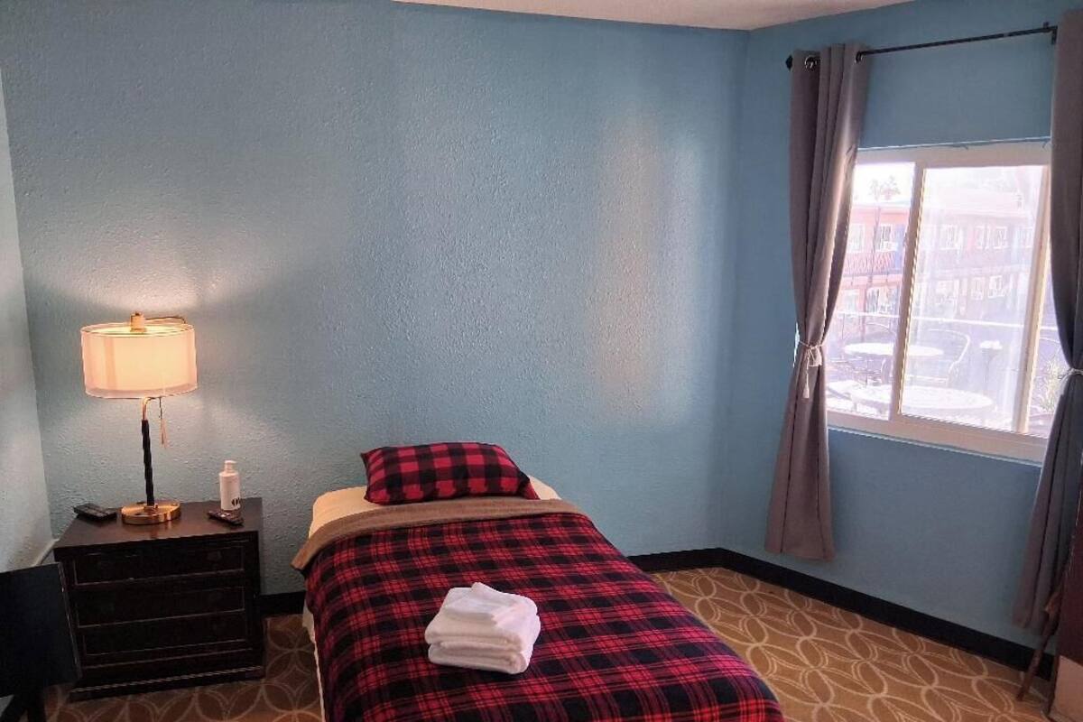 Une petite chambre d’hôtel avec un lit simple, une table de chevet avec une lampe et une fenêtre avec des rideaux.