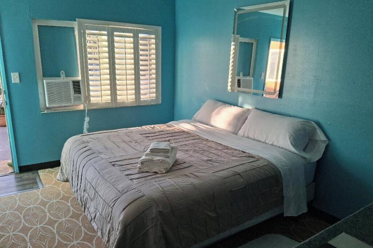 Une chambre à coucher avec un lit, une fenêtre avec des stores, un miroir et une petite table de chevet.