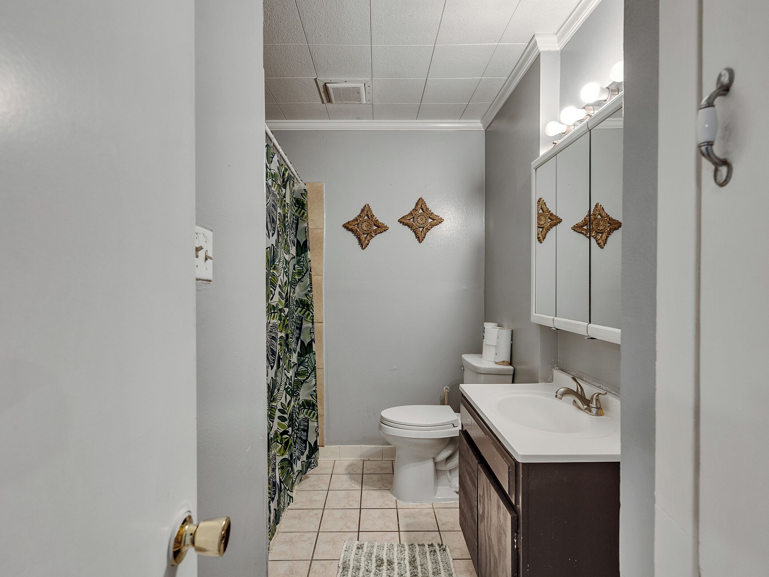 Une salle de bain avec des toilettes, un lavabo et un miroir. Les murs sont décorés de carreaux et il y a un rideau de douche orné d’un motif de feuilles.