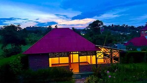 A house with a red roof and a well-lit interior, surrounded by greenery and a clear sky. - Lights of Kazinga Homestay (Kichwamba)