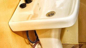 A white pedestal sink with a black faucet, a visible drain, and a flexible hose connected to the wall. - Lights of Kazinga Homestay (Kichwamba)