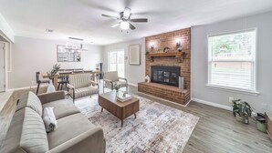A living room with a fireplace, a ceiling fan, and a dining area with chairs and a table. - 3-bed house in Denton, pool, games, firepit in a huge private fenced yard (Denton)