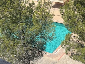Ein klares, blaues Schwimmbecken, umgeben von Bäumen, mit einer Terrassenanlage und einem Gebäude im Hintergrund.
