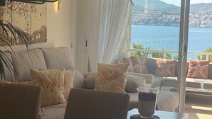 Een woonkamer met een bank, stoelen en een glazen tafel met een drankje. Een groot raam biedt uitzicht op een waterpartij en een stad in de verte.