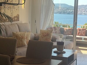 Ein Wohnzimmer mit einer Couch, Sesseln und einem Glastisch mit einem Getränk. Ein großes Fenster bietet Blick auf ein Gewässer und eine ferne Stadt.