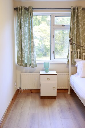 Quarto com janela, mesa de cabeceira com abajur, radiador e cama.