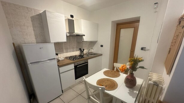 A compact kitchen with white cabinets, a white refrigerator, a stove, and a sink. There's a dining table with two chairs and a vase with flowers. - Comfort in the heart of Bari (Bari)