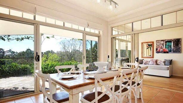Un coin repas avec une table en bois et des chaises blanches, de grandes fenêtres donnant sur un paysage verdoyant, et un canapé avec des coussins.