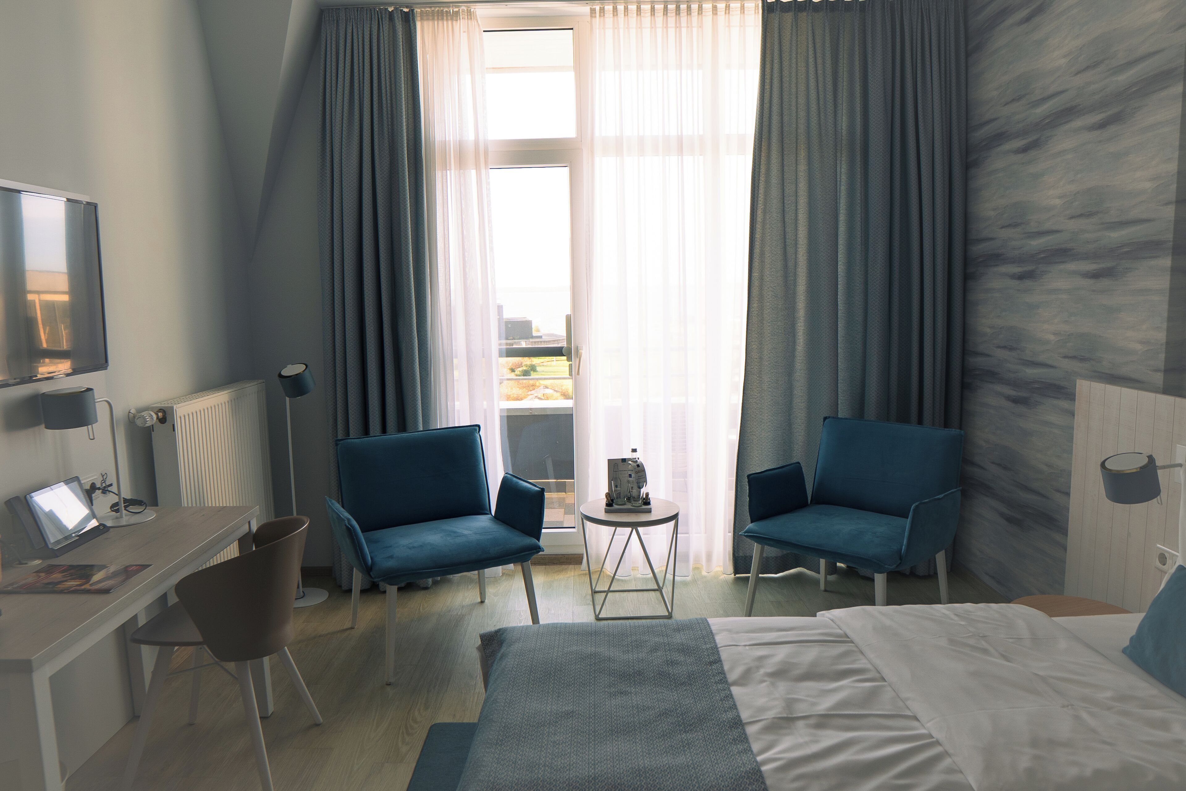 A hotel room with a bed, two blue armchairs, a small table, a desk with a laptop, and a view of the outdoors through sheer curtains.