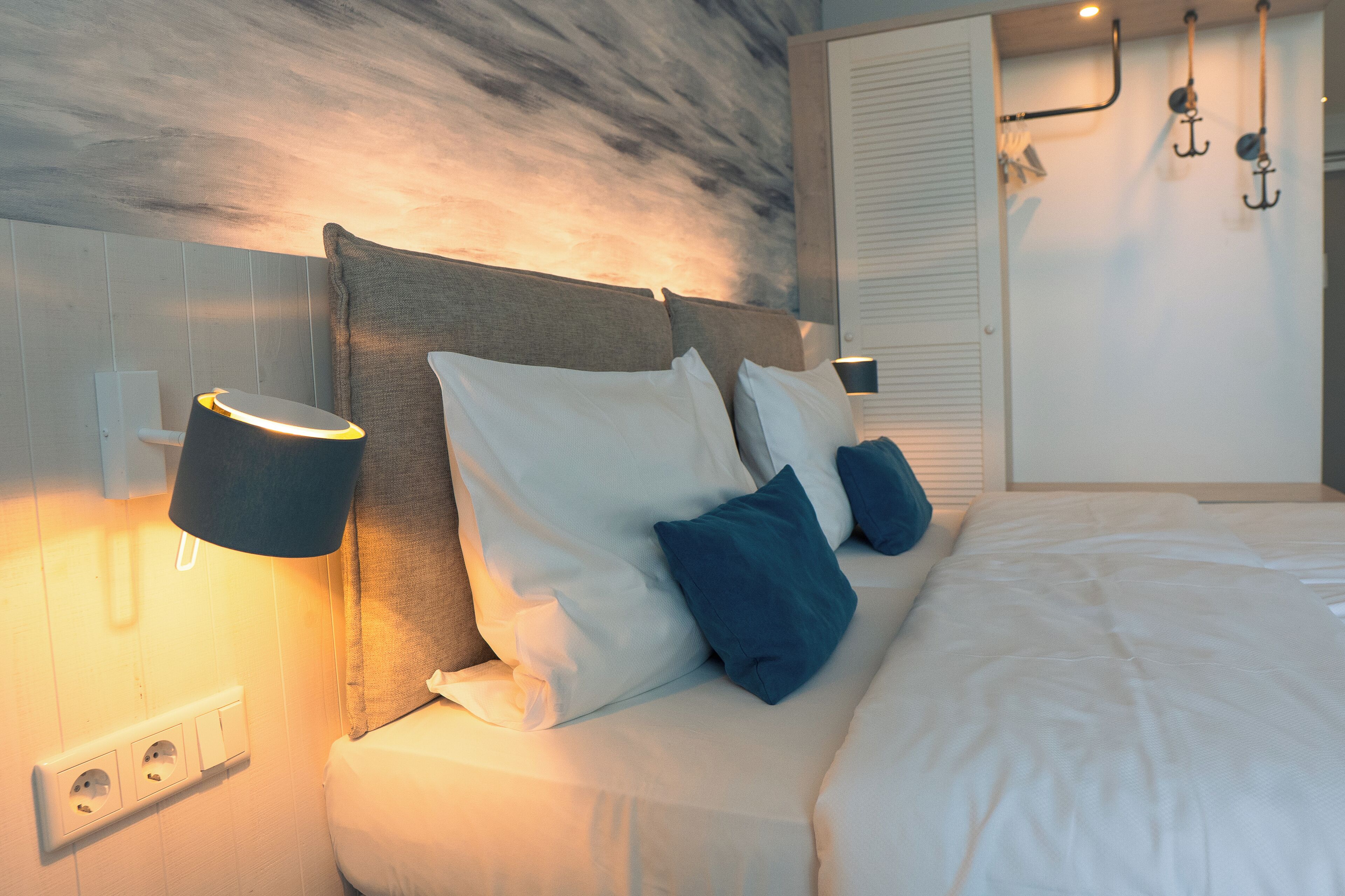 Ein ordentlich bezogenes Bett mit weißer und blauer Bettwäsche, ein Kopfteil mit einer Lampe und eine wandmontierte Steckdose.