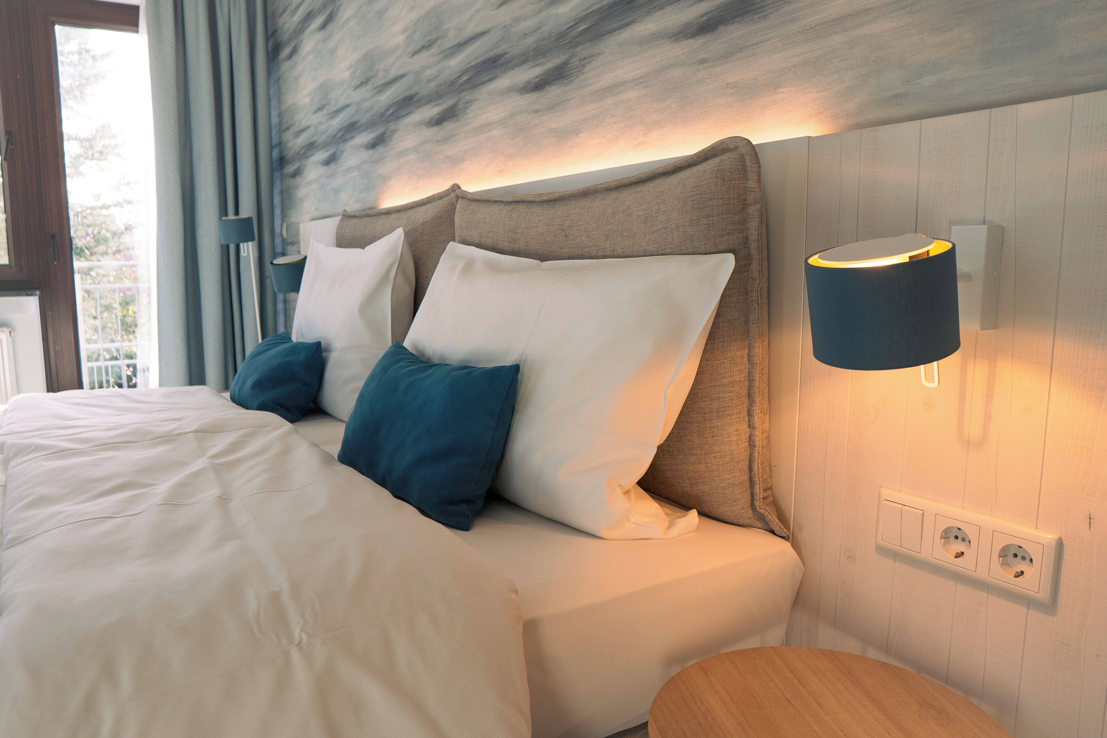 Una cama bien hecha con almohadas blancas y azules, una mesita de noche de madera y una lámpara de pared.
