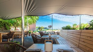 Un área de comedor al aire libre con mesas y sillas de madera, un objeto cilíndrico blanco sobre una mesa, y una vista de la ciudad al fondo.